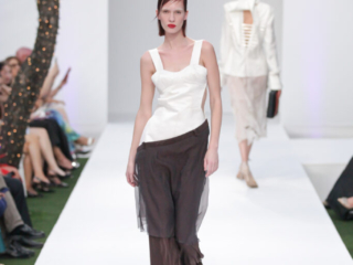 Modella con top bianco asimmetrico e pantaloni marroni trasparenti.